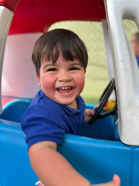 A toddler boy grinning behind the wheel of a toy car